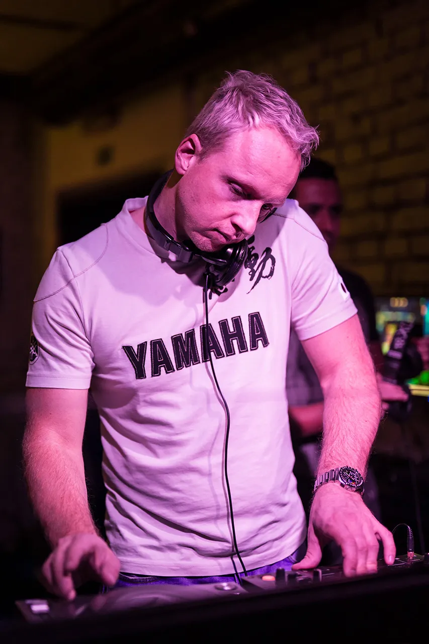 Muž, Béla En, stojící za mixážním pultem a svírající sluchátka hlavou a ramenem, soustředící se na svůj mix v barevně nasvíceném klubu