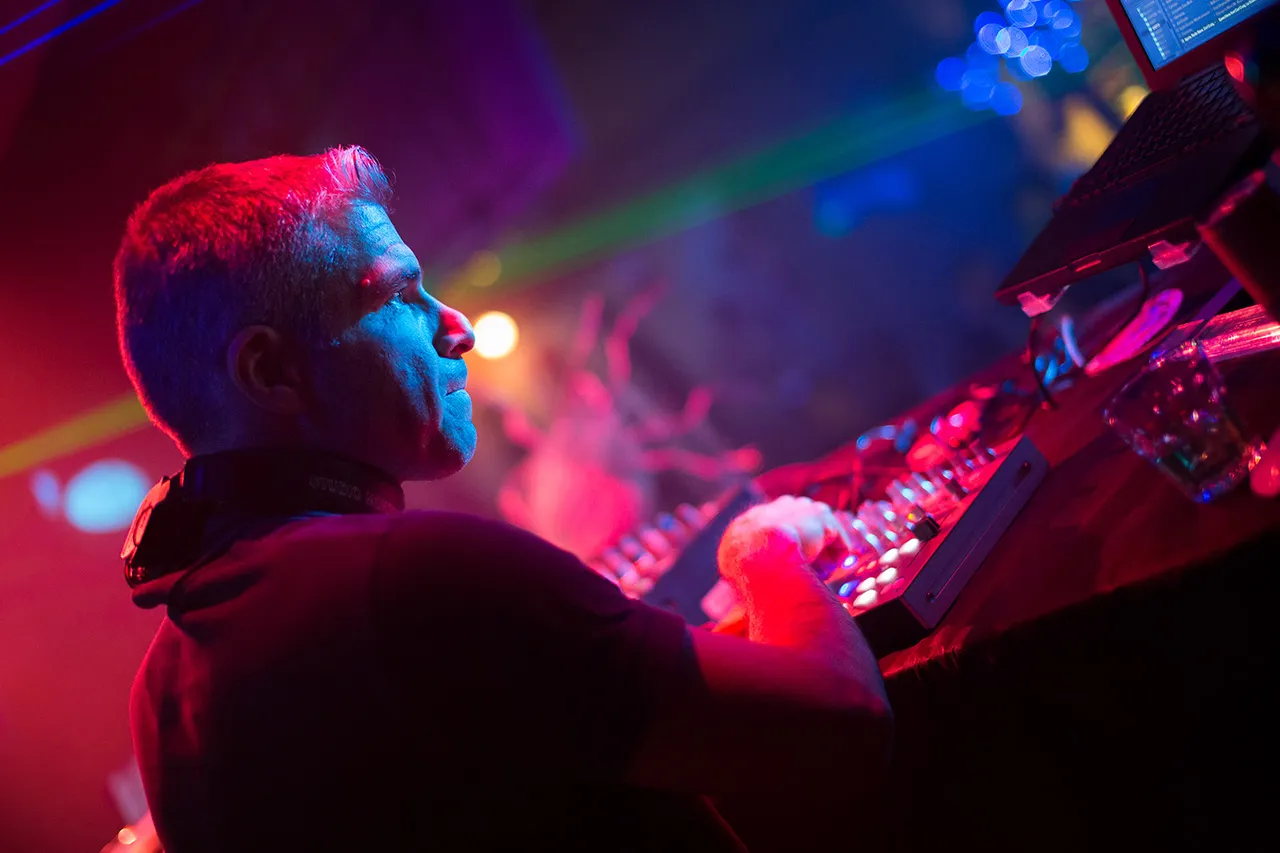 Muž, Marco Zaffarano, stojící za mixážním pultem, soustředící se na svůj mix v barevně nasvíceném klubu
