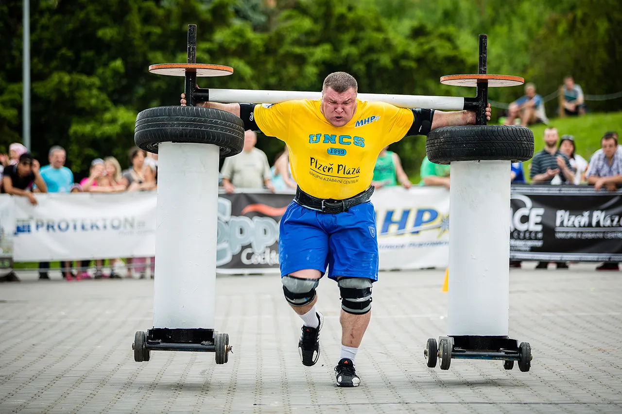 Mohutný závodník strongman ve žlutém tričku UNCS a modrých šortkách kráčí s obrovskou konstrukcí na ramenou, na které jsou upevněny pneumatiky. Snímek zachycuje extrémní fyzické vypětí a soustředění při disciplíně Yoke Walk před diváky