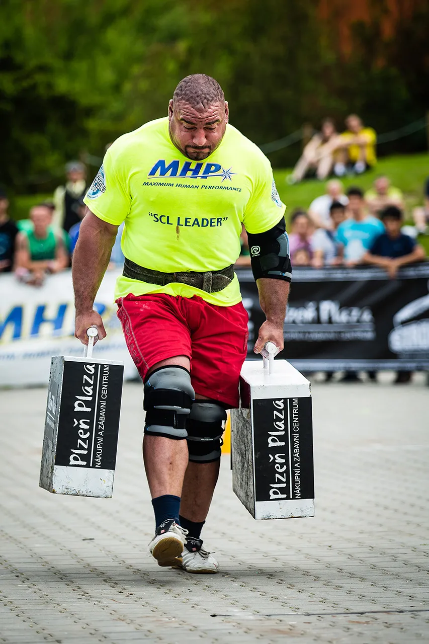 Závodník strongman v plném nasazení při disciplíně Farmer's Walk (Farmářská chůze) s těžkými kufry v obou rukách.