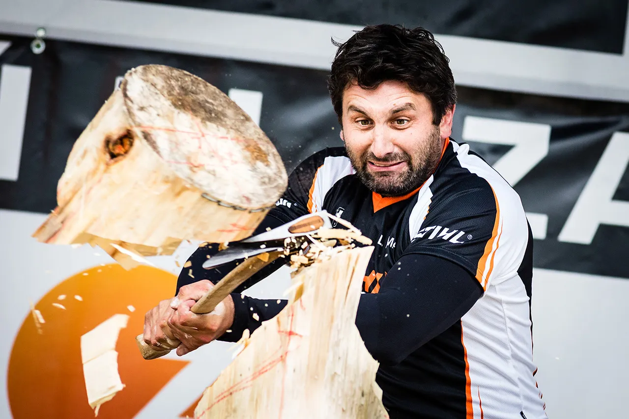 Závodník v dresu STIHL Timbersports dokončuje disciplínu Standing Block Chop. Fotografie zachycuje závodníka v momentě, kdy finálním úderem sekyry odsekává horní část kmene.
