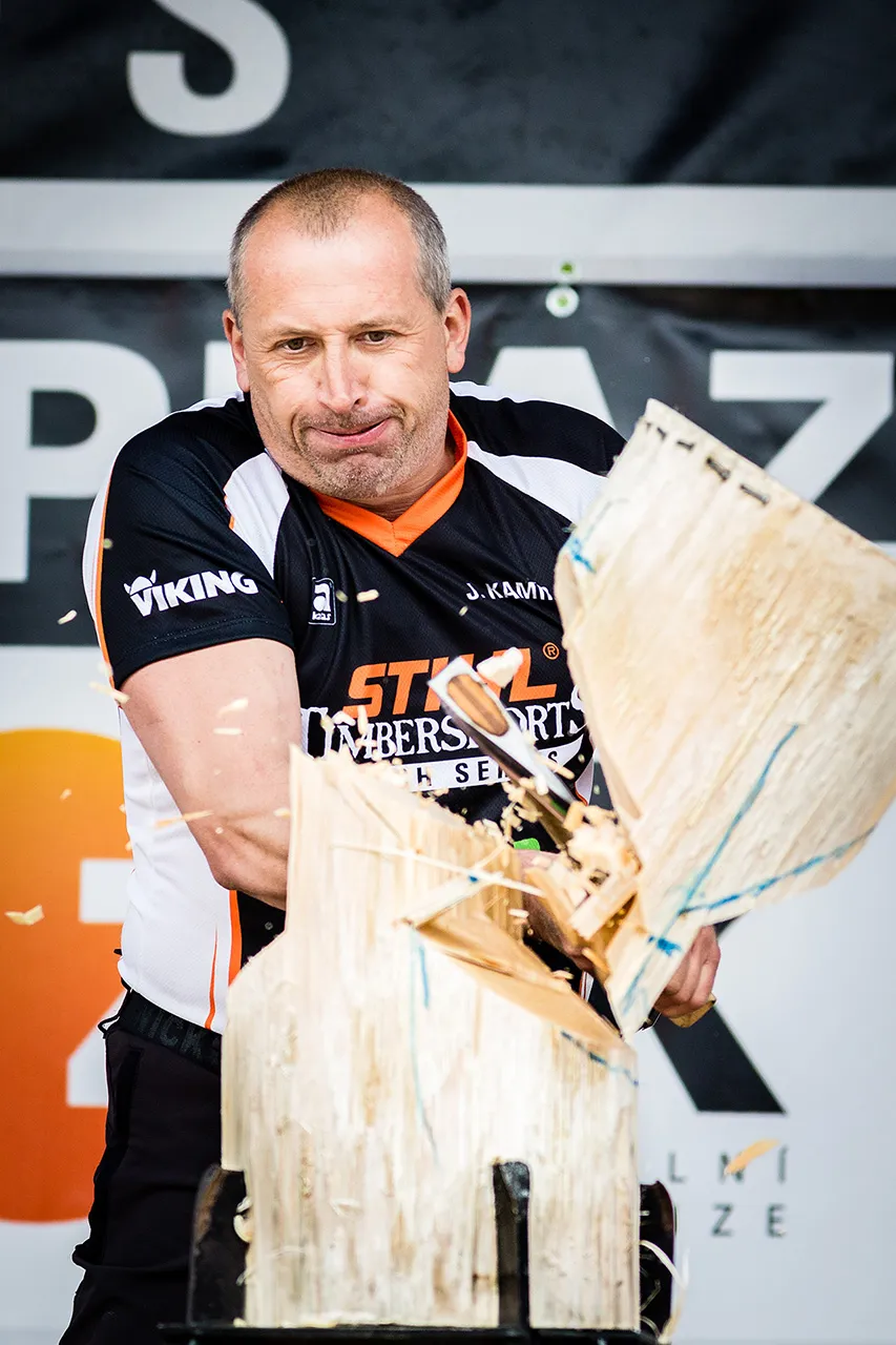 Závodník STIHL Timbersports dokončuje sek v disciplíně Standing Block Chop; detailní pohled zachycuje třísky a rozpolcený kmen v okamžiku nárazu sekery.