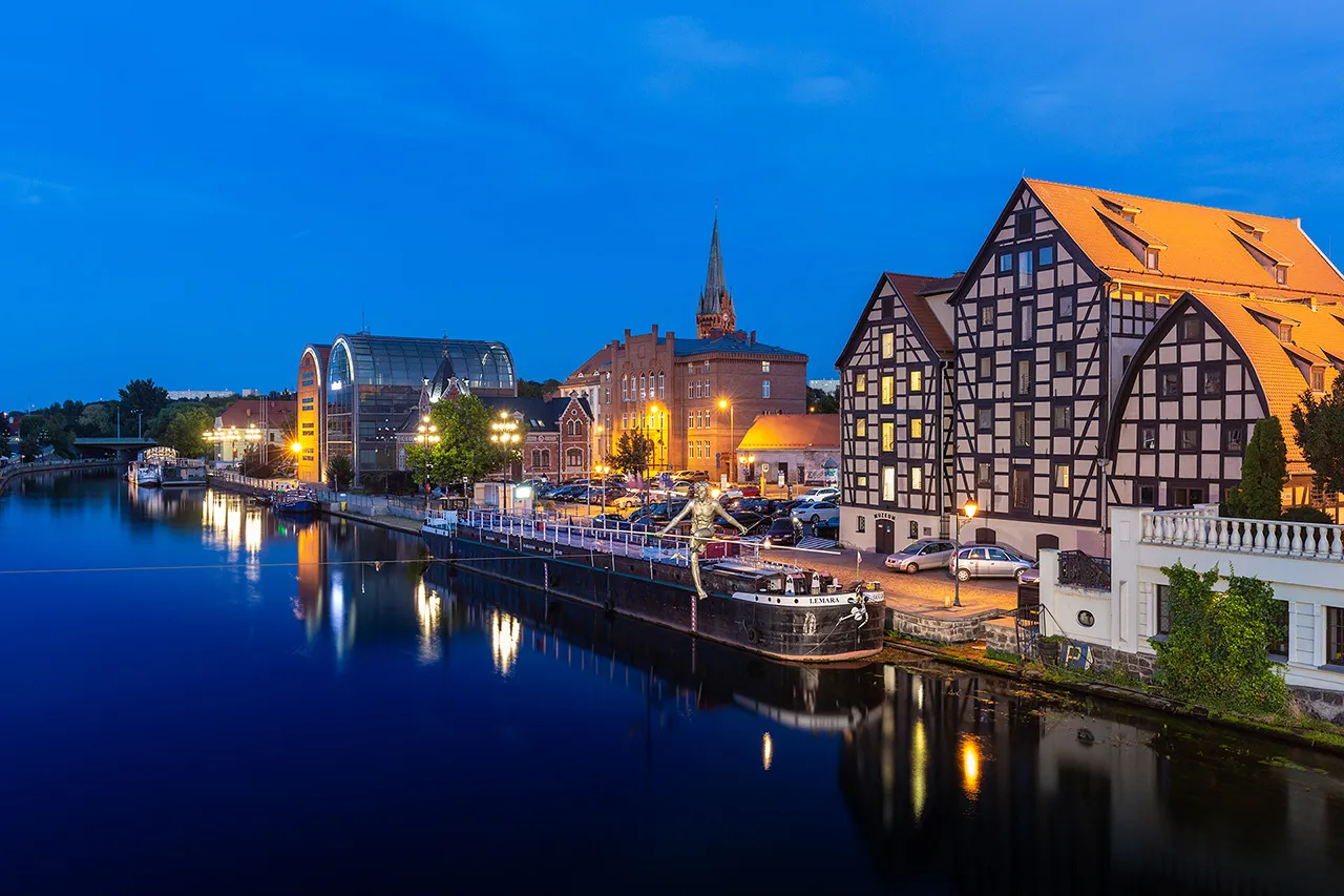Historické sýpky a moderní budovy na nábřeží řeky Brda v Bydhošti během modré hodinky.