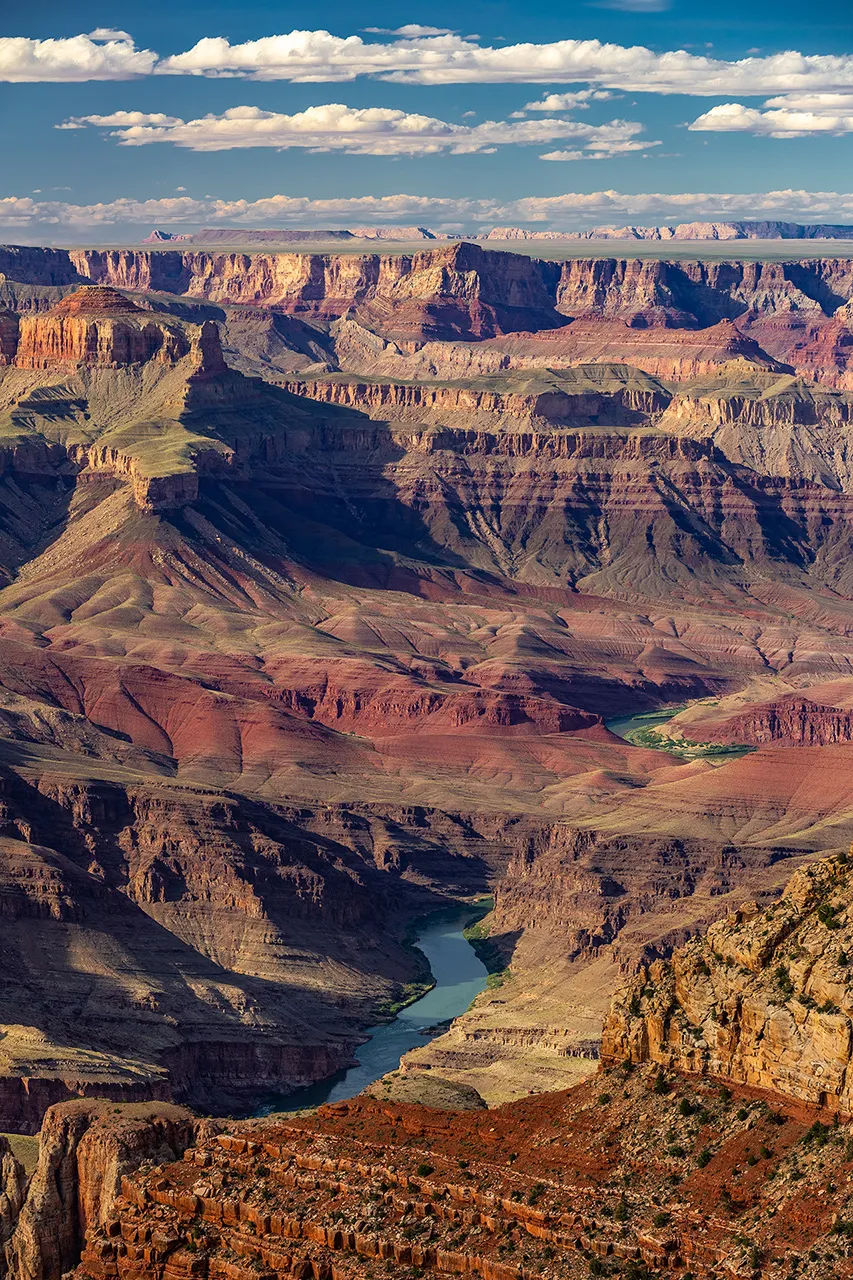 Pohled do hloubi Grand Canyonu na klikatící se řeku Colorado obklopenou vrstvenými skalními útvary pod letní oblohou.