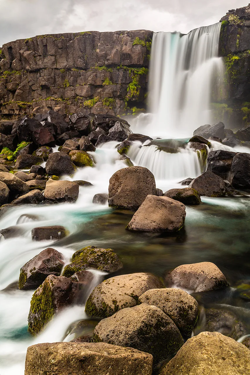 Dlouhá expozice vodopádu Öxarárfoss v národním parku Þingvellir, kde voda stéká přes tmavé balvany v tektonické trhlině.